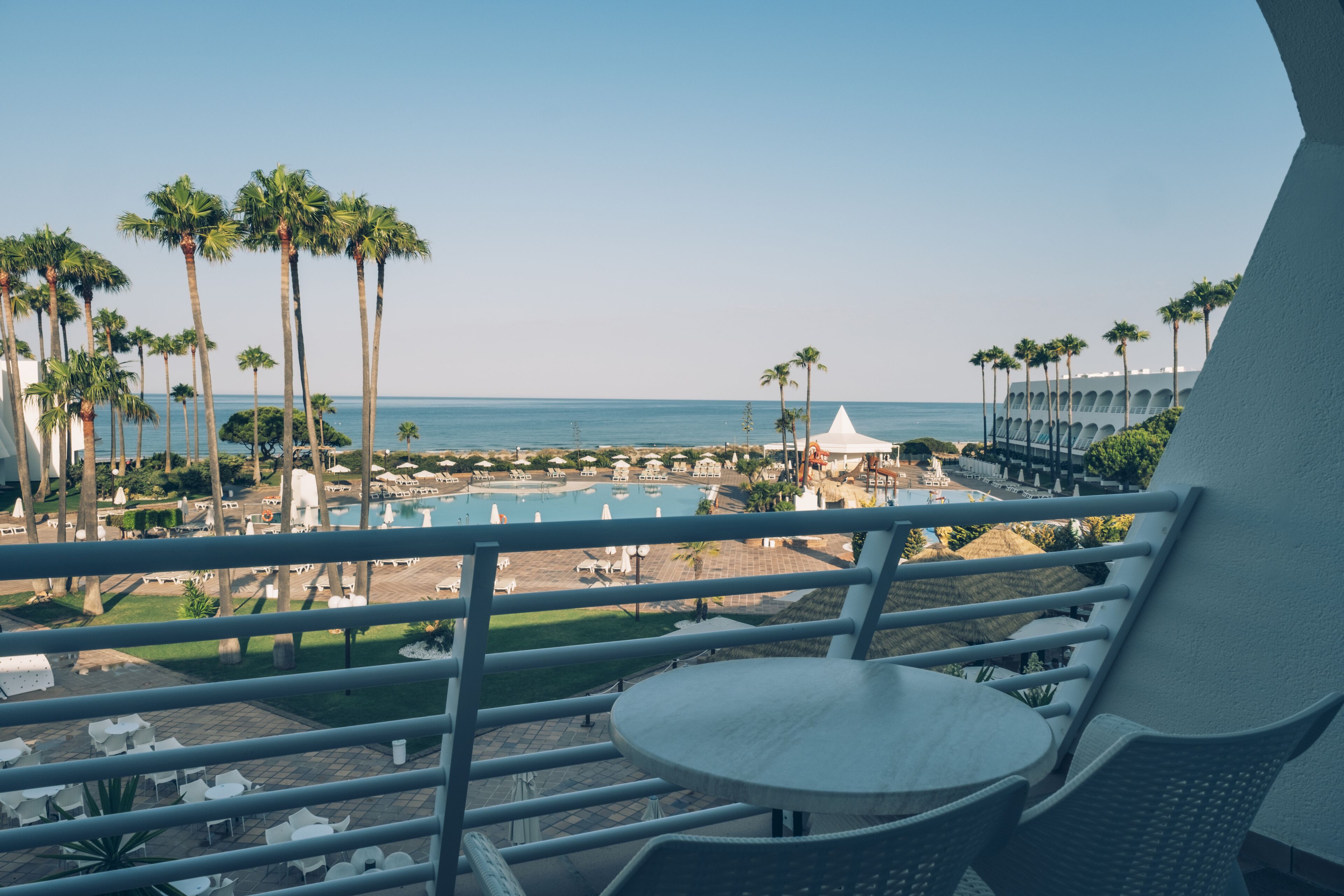 Un balcón con vista a una piscina, palmeras y una playa.