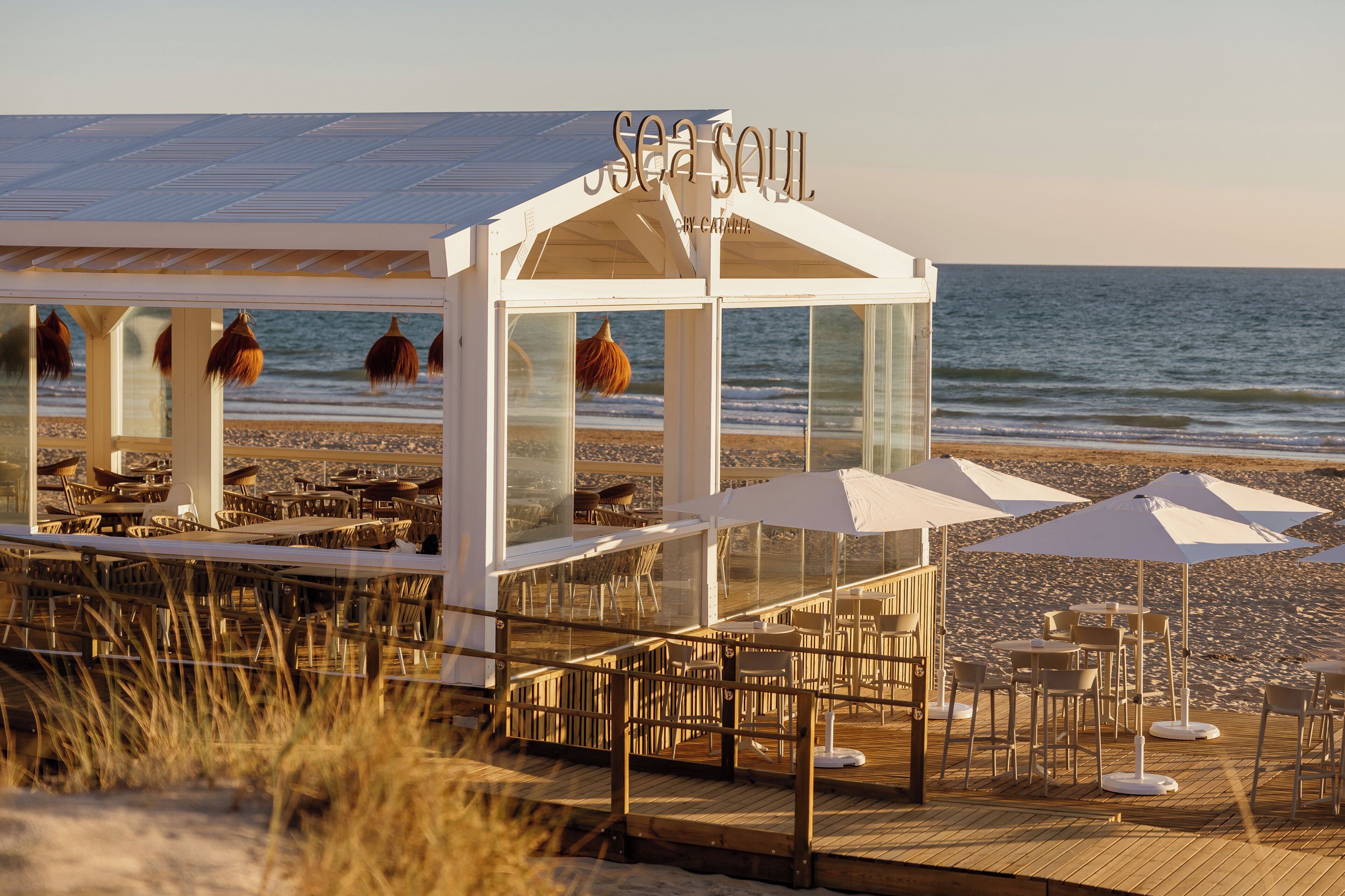 A beachside restaurant with outdoor seating, a glass enclosure, and a view of the sea.