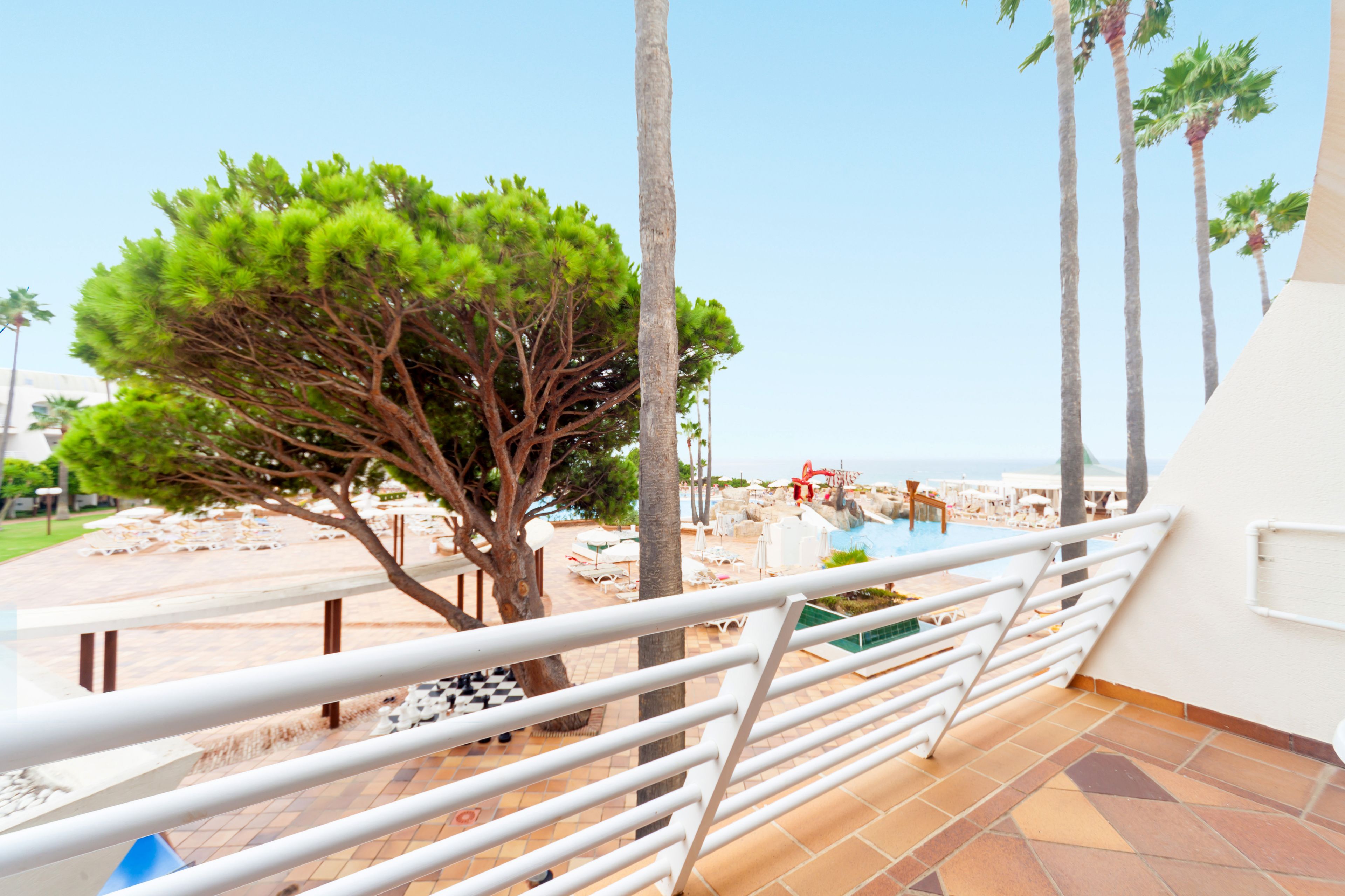 Un balcón con vista a la piscina, palmeras y mobiliario exterior.
