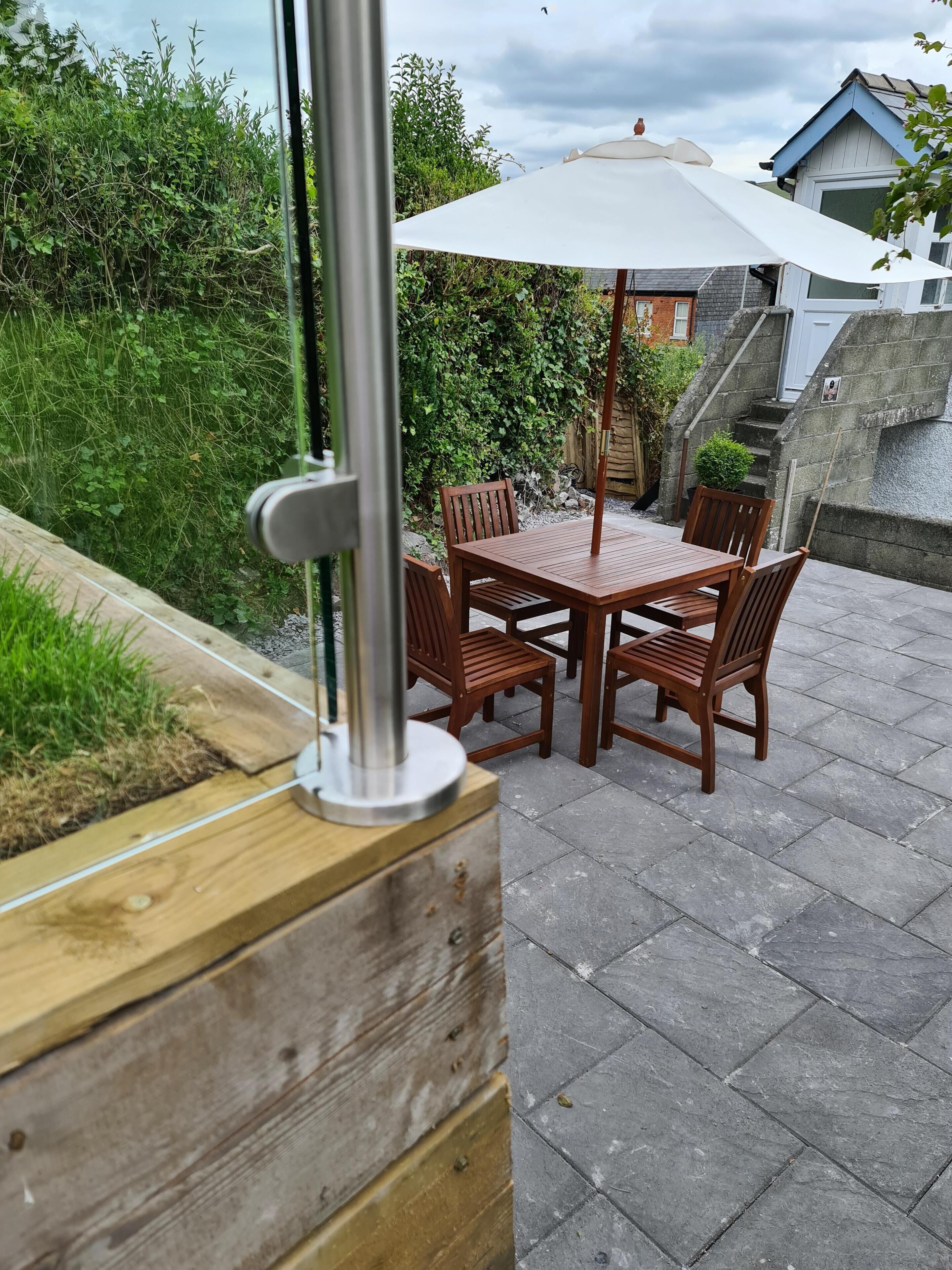 Een terras met een houten tafel en stoelen, een witte parasol en een metalen paal.