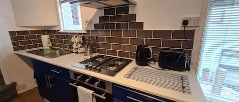 A modern kitchen with a gas stove, a kettle, and a toaster. There is a window with blinds, a sink, and a countertop with a dish rack.