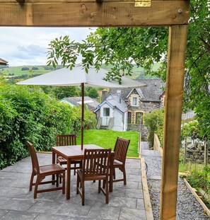 A wooden pergola with a table and chairs, an umbrella, and a view of a stone house with a garden. - Ty Morgans Apartments (Rhayader)