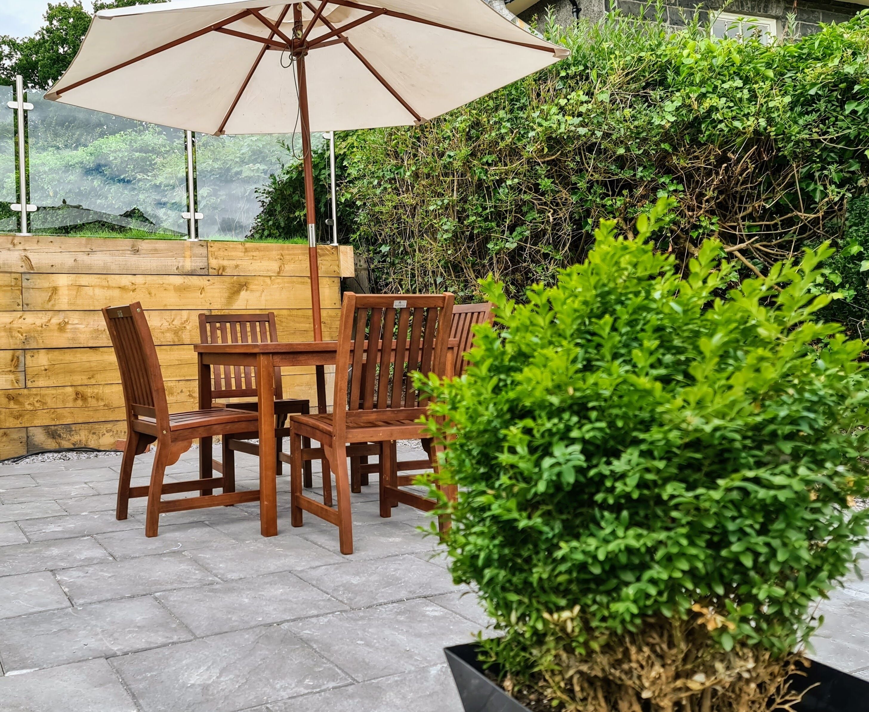 A patio area with a wooden table and chairs, a beige umbrella, and a potted plant.