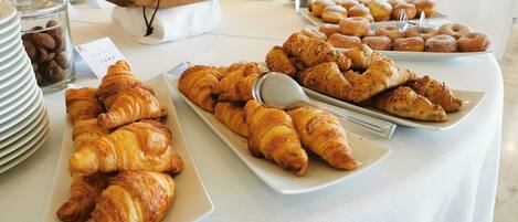 Una mesa de buffet con diversos pasteles, entre ellos cruasanes y rosquillas, y un tarro de chispas de chocolate.