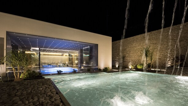 Un área de piscina moderna al aire libre con un spa, un muro de piedra y un edificio con grandes ventanales.