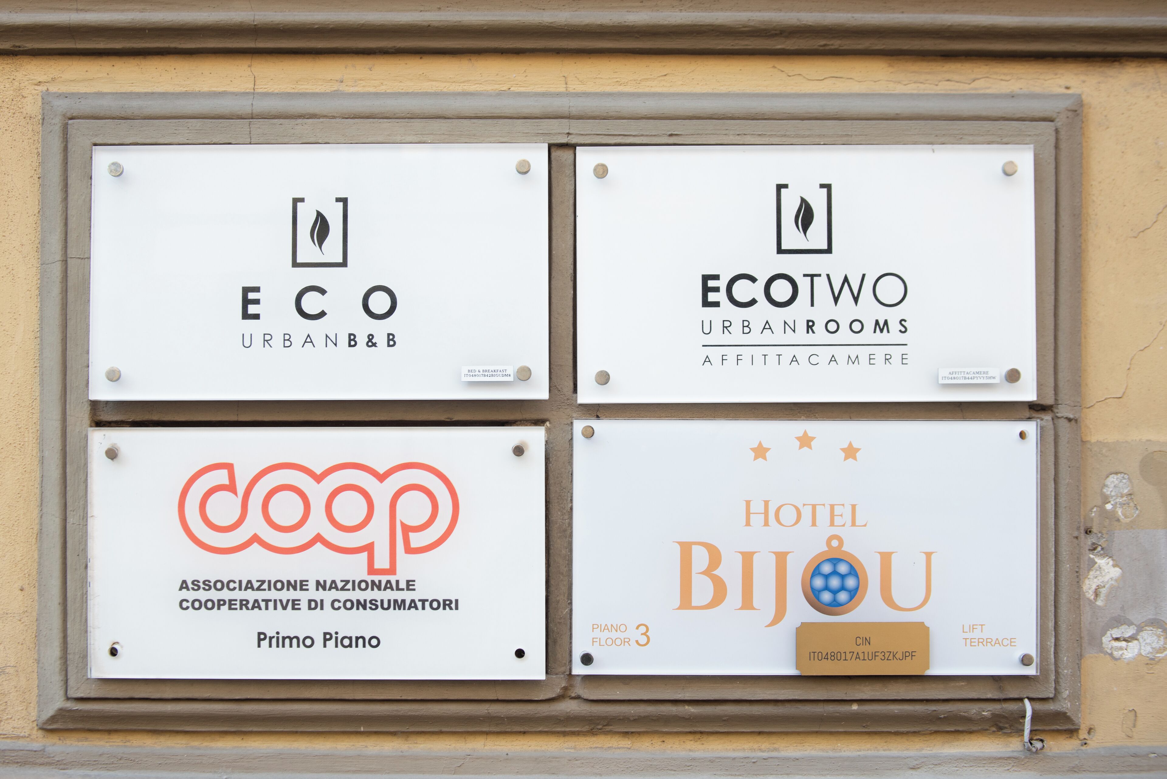 Four signs mounted on a wall, each representing different entities: ECO URBAN&B&B, ECOTWO URBANROOMS AFFITTACAMERE, COOP ASSOCIAZIONE NAZIONALE COOPERATIVE DI CONSUMATORI Primo Piano, and HOTEL BIJOU Piano Floor 3 Lift Terrace.