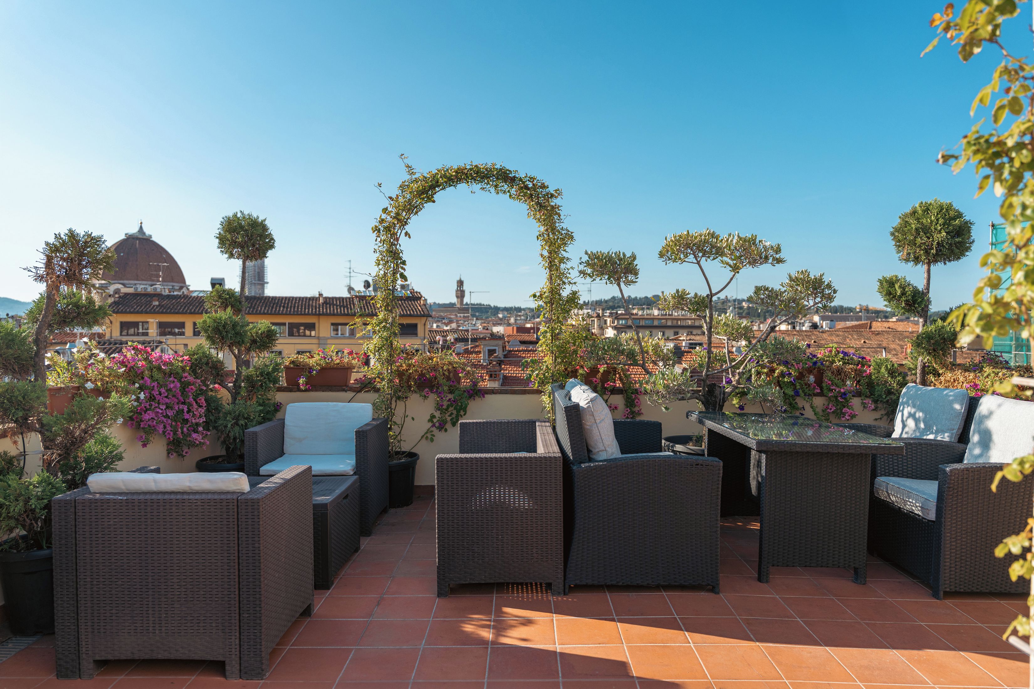 A rooftop terrace with wicker furniture, potted plants, and a view of the cityscape.