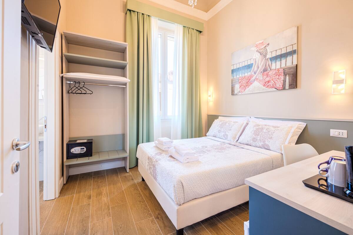 A hotel room with a bed, a desk, a wardrobe, and a painting on the wall.