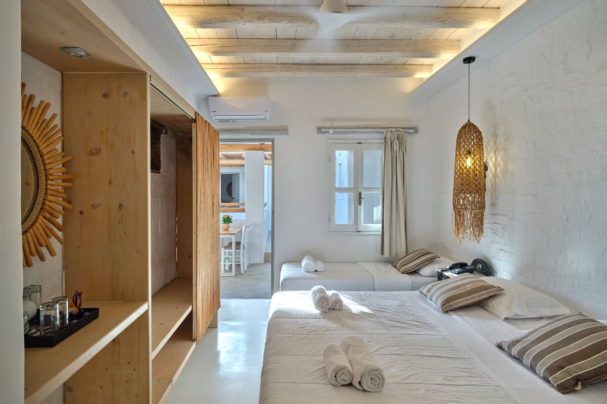 A modern bedroom with a wooden ceiling, a large bed with rolled towels, a hanging light fixture, and a view of a dining area.