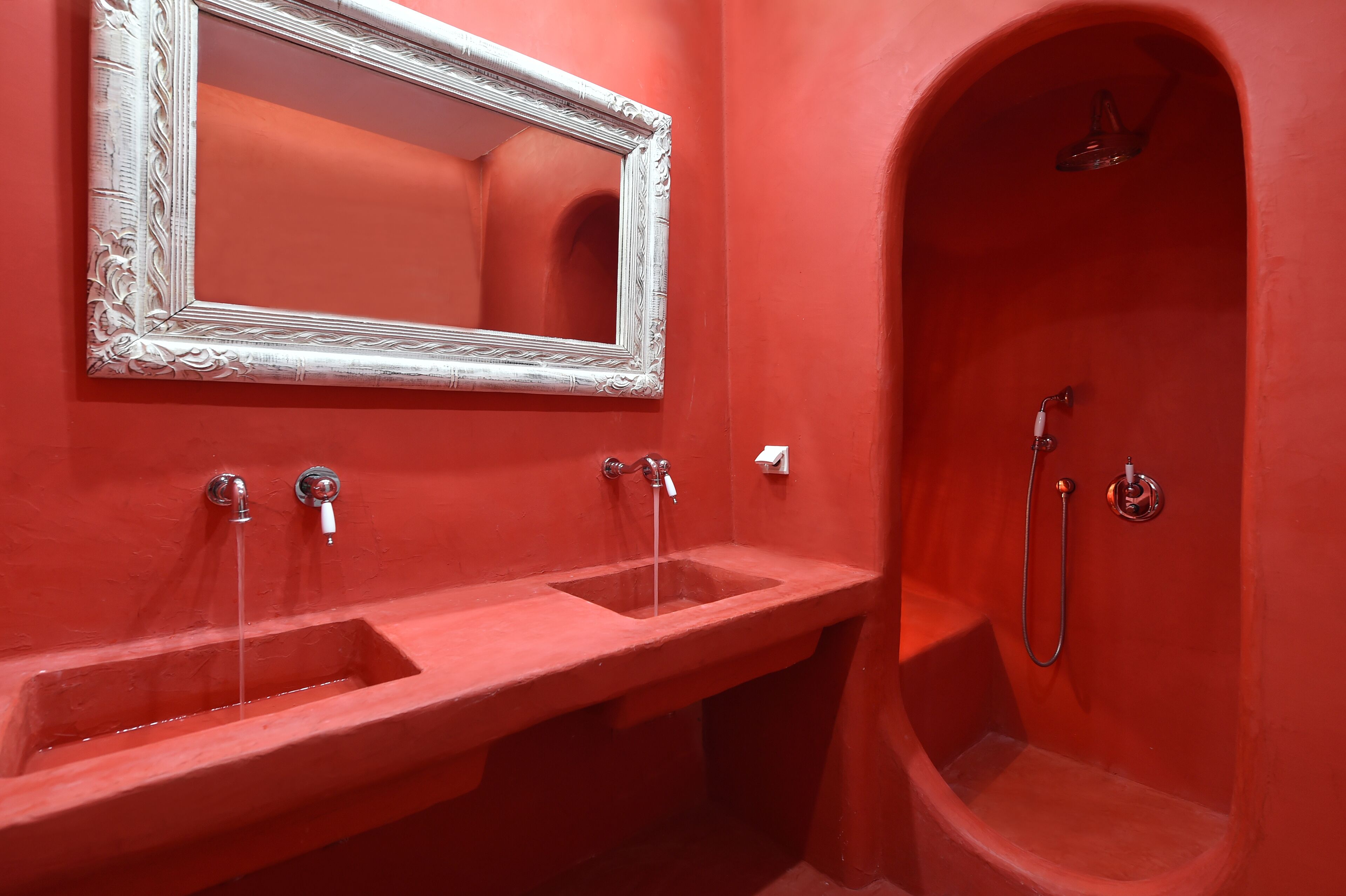 A bathroom with a red wall, a white ornate mirror, two sinks, and a shower area.
