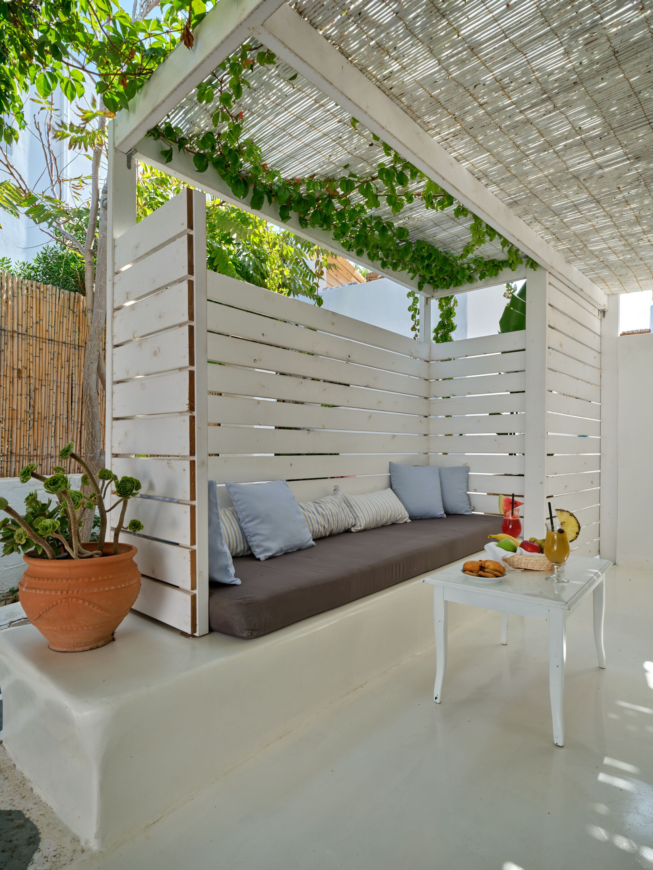 A white outdoor seating area with a cushioned bench, a white table, and a potted plant.