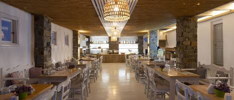 A spacious restaurant with wooden tables and chairs, a central chandelier, stone walls, and a bar area in the background.