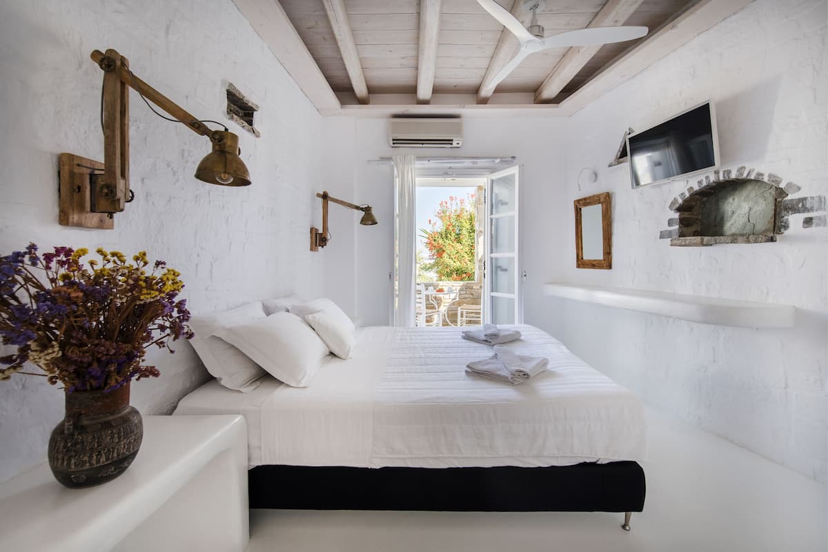 A bedroom with a bed, a wooden desk, a vase with flowers, a fireplace, and a TV mounted on the wall.