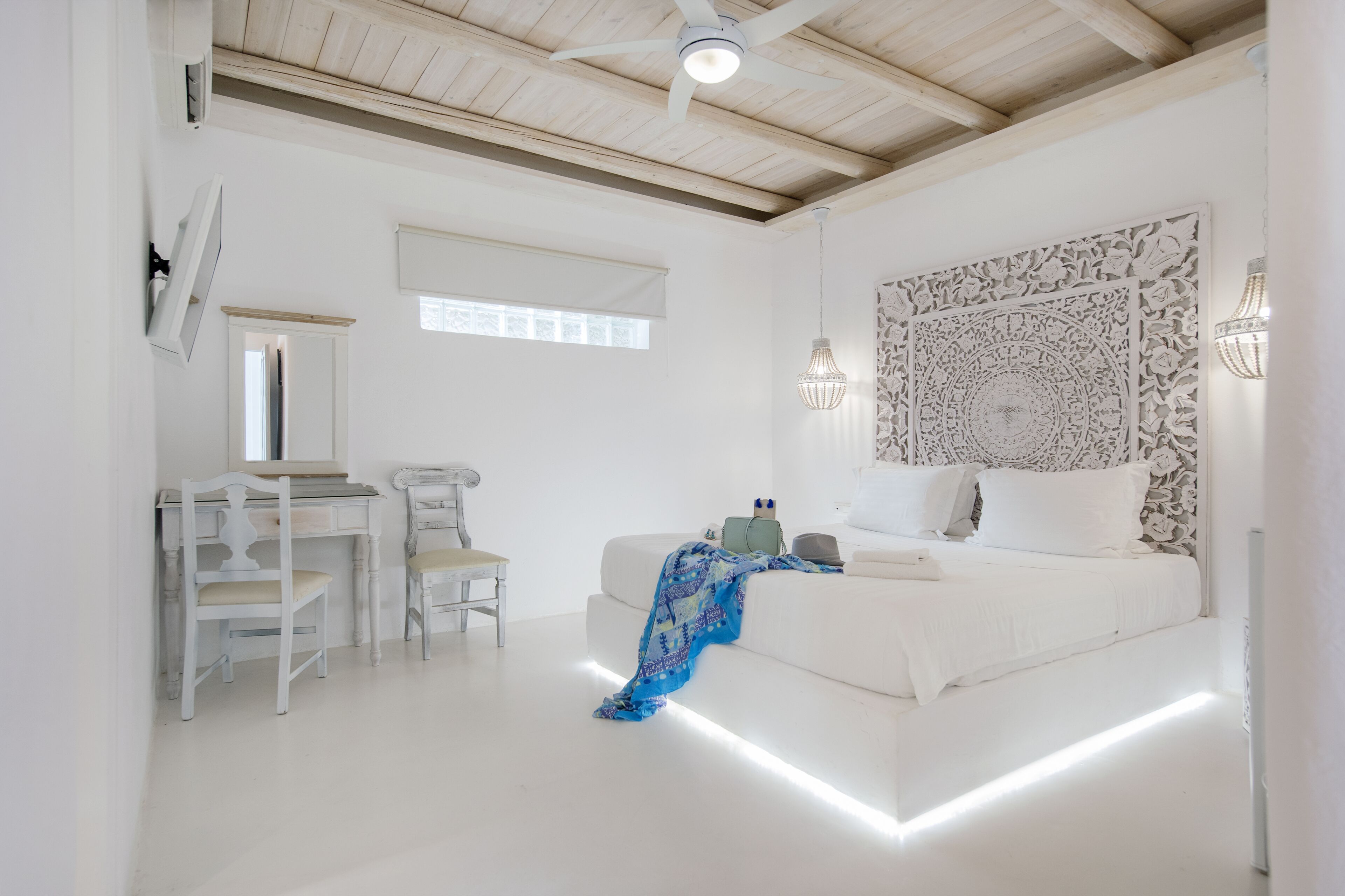 A bedroom with a bed, a desk, two chairs, a ceiling fan, and a decorative wall panel.