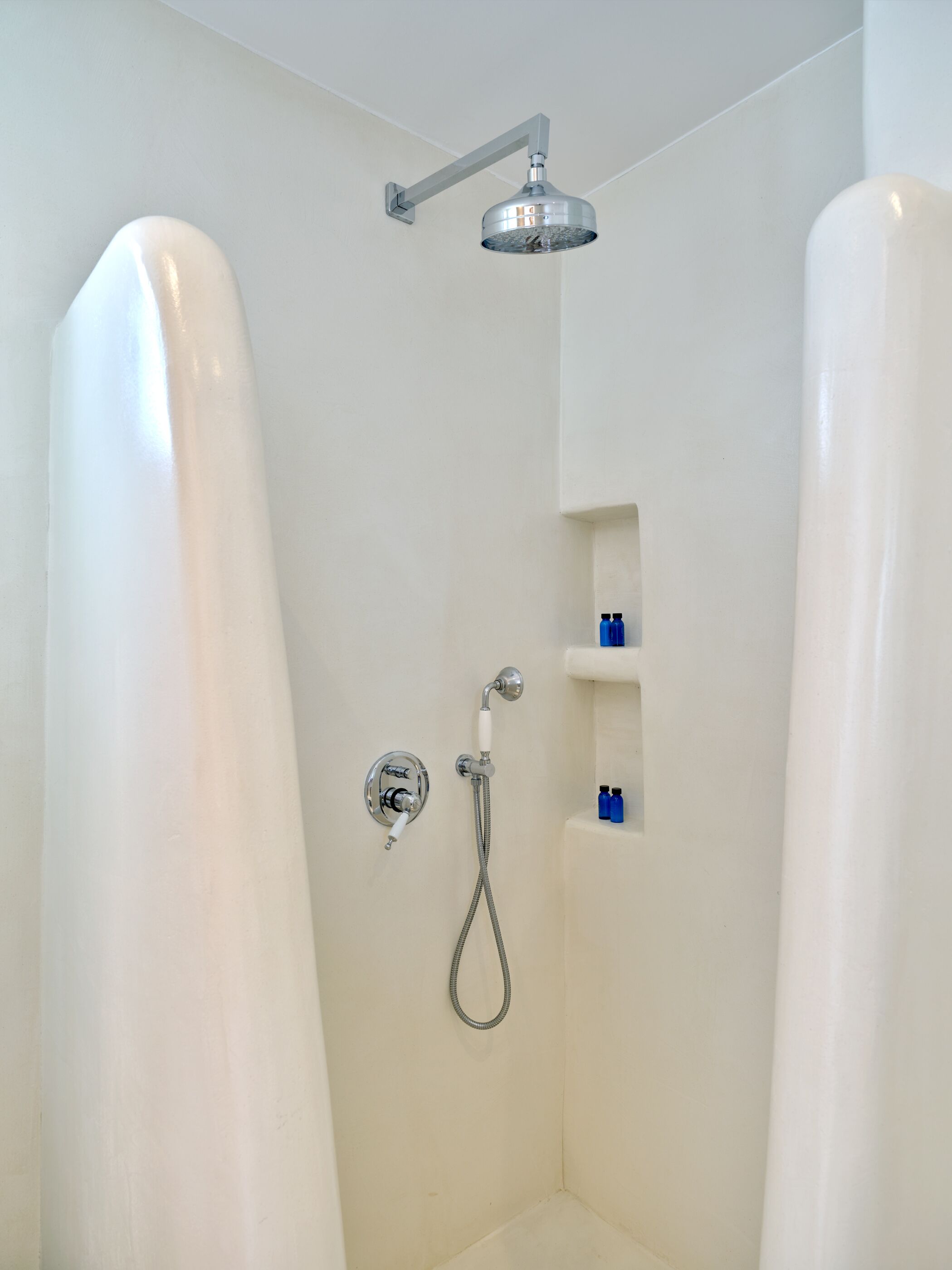 A shower with a handheld showerhead and a wall-mounted faucet. There are two small blue bottles on a shelf.