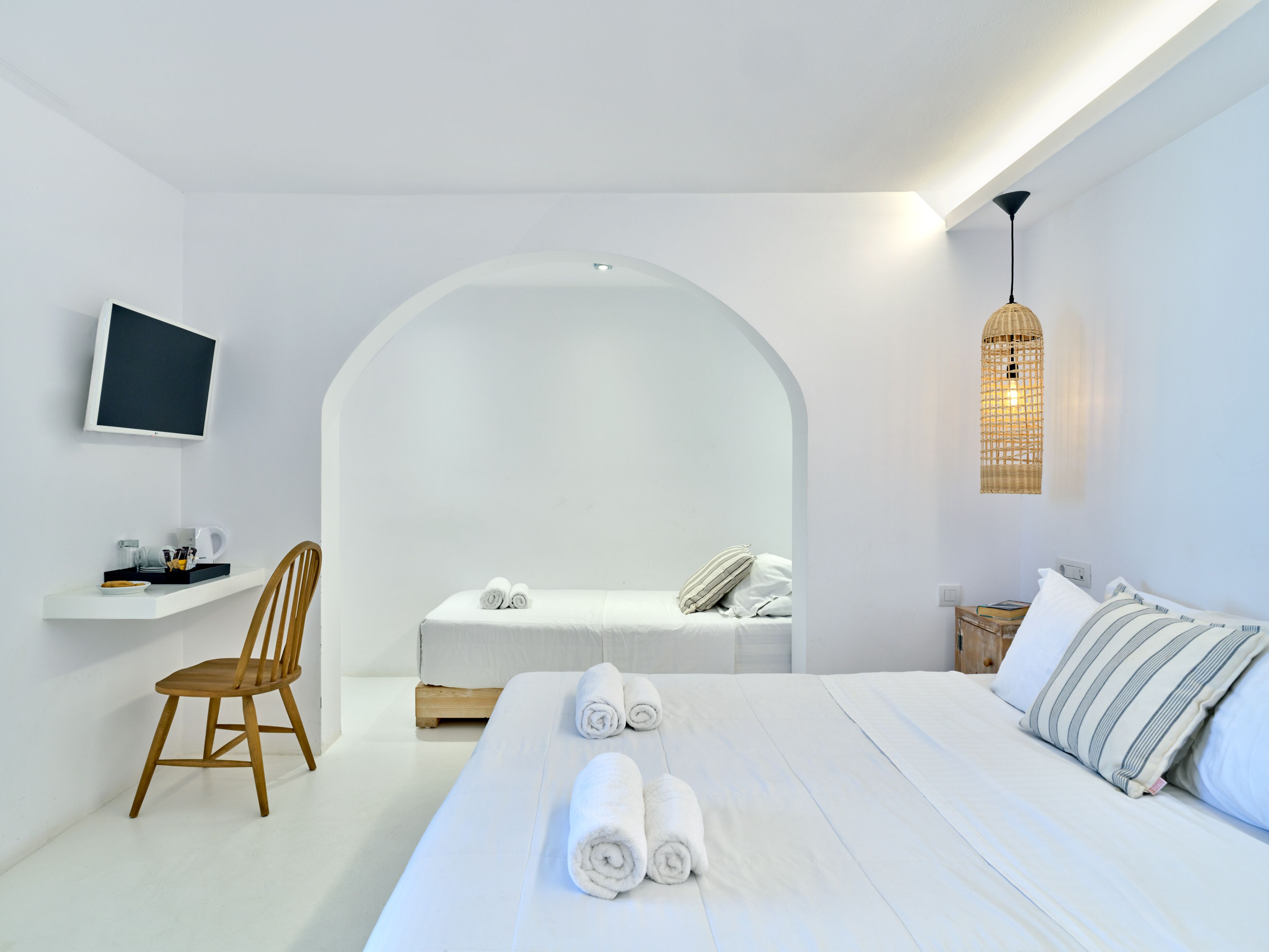 A minimalist hotel room with a bed, a chair, a small table with a coffee maker, and a television on the wall.