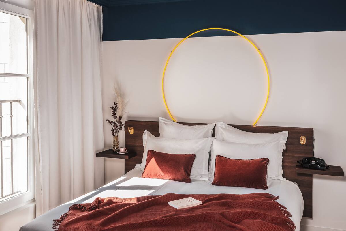 A neatly made bed with white and red bedding, a bedside table with a lamp, and a wall-mounted circular light fixture.