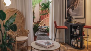 A cozy room with a wicker chair, a round table with a book, a bar area with bottles, and a view of a garden through a doorway. - Seven Hotel Paris (Paris)