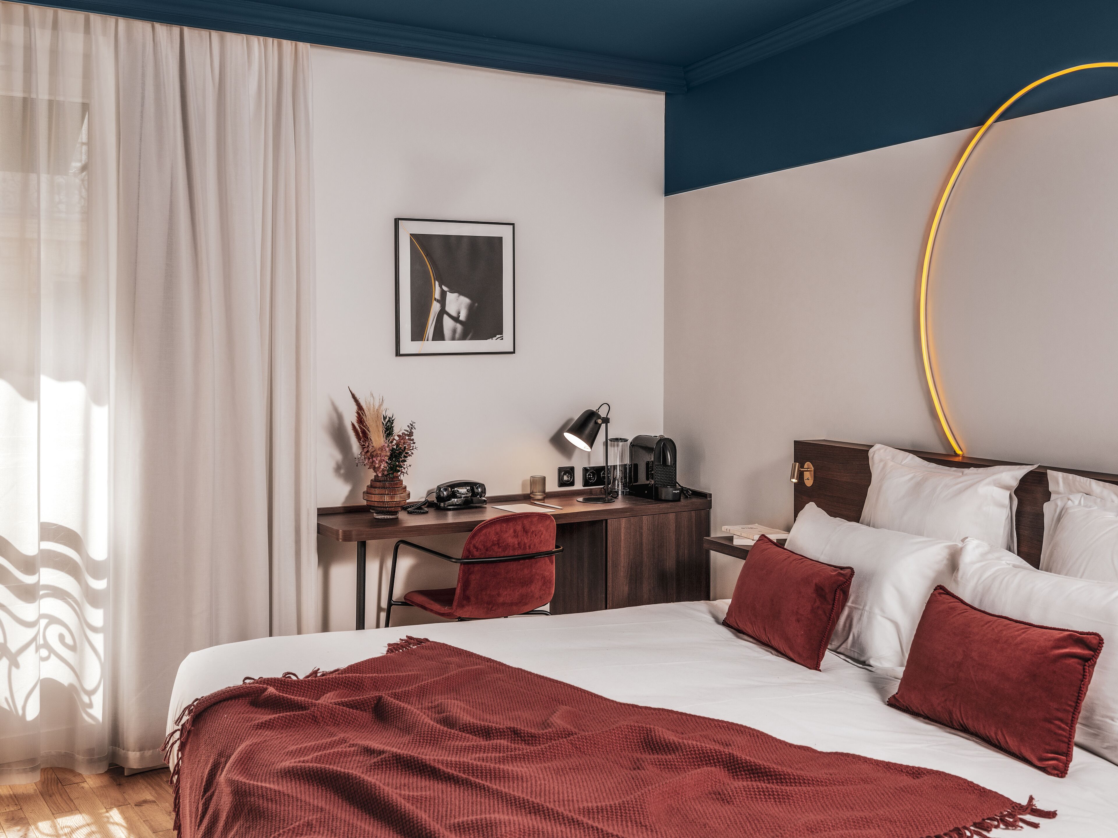 A hotel room with a bed, a desk with a lamp, a coffee maker, and a framed picture on the wall.
