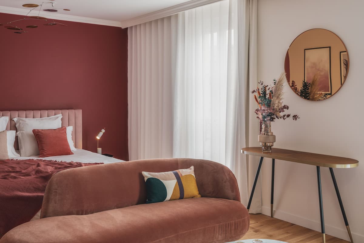 A modern bedroom with a red accent wall, a tufted headboard, a sofa with colorful cushions, a round coffee table, and a bedside lamp.