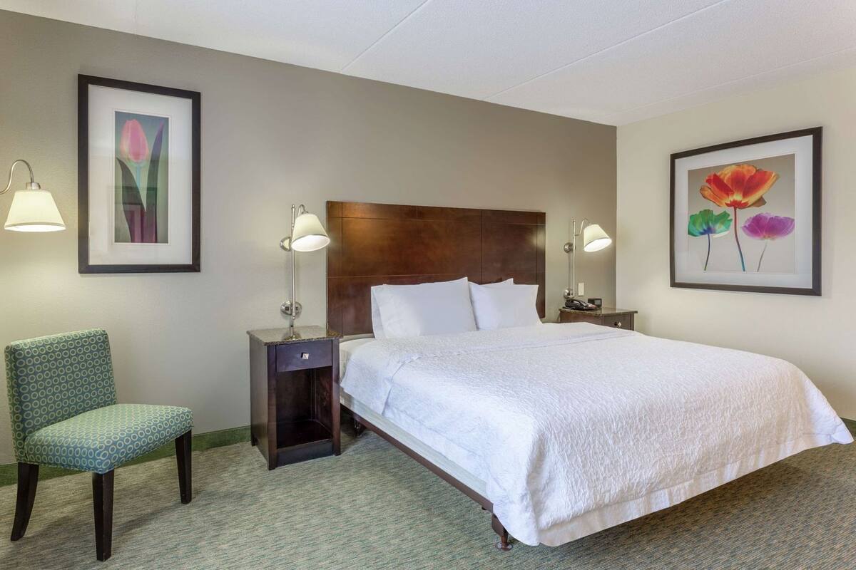 A hotel room with a large bed, two bedside tables with lamps, a chair, and framed floral artwork on the walls.