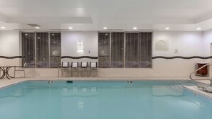 Ein Swimmingpool mit glasklarer, blauer Wasseroberfläche, umgeben von einer gefliesten Liegefläche. Auf beiden Seiten des Pools befinden sich jeweils zwei Fensterreihen mit Rollläden.