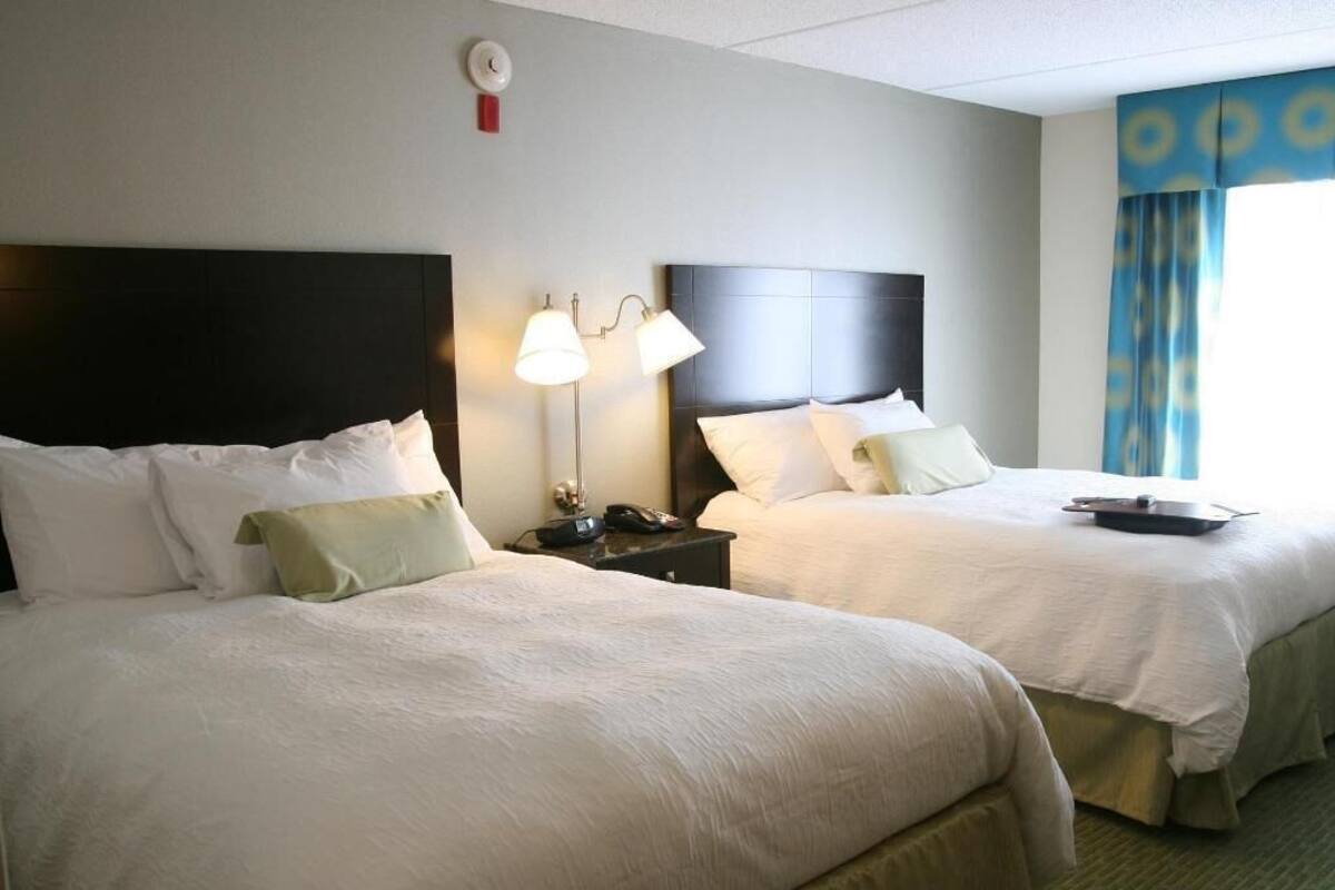A hotel room with two beds, a nightstand with a lamp, and a window with curtains.