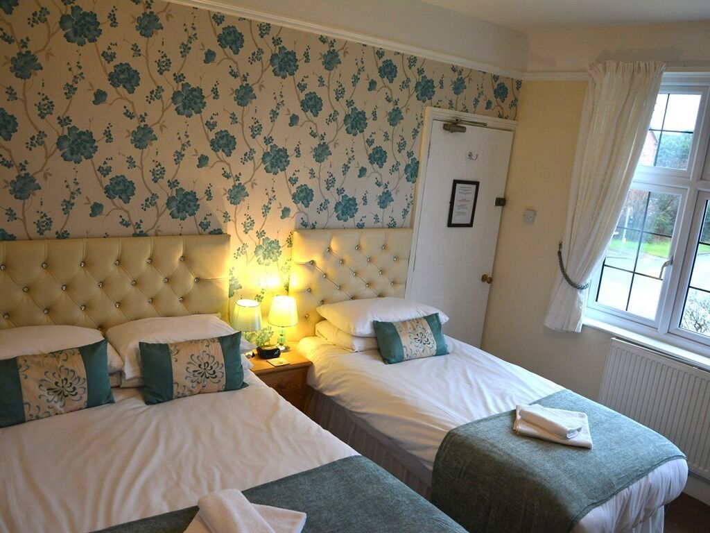 Une chambre d’hôtel avec deux lits, un papier peint à motifs floraux, une tête de lit capitonnée, une table de chevet avec une lampe et une fenêtre avec des rideaux.