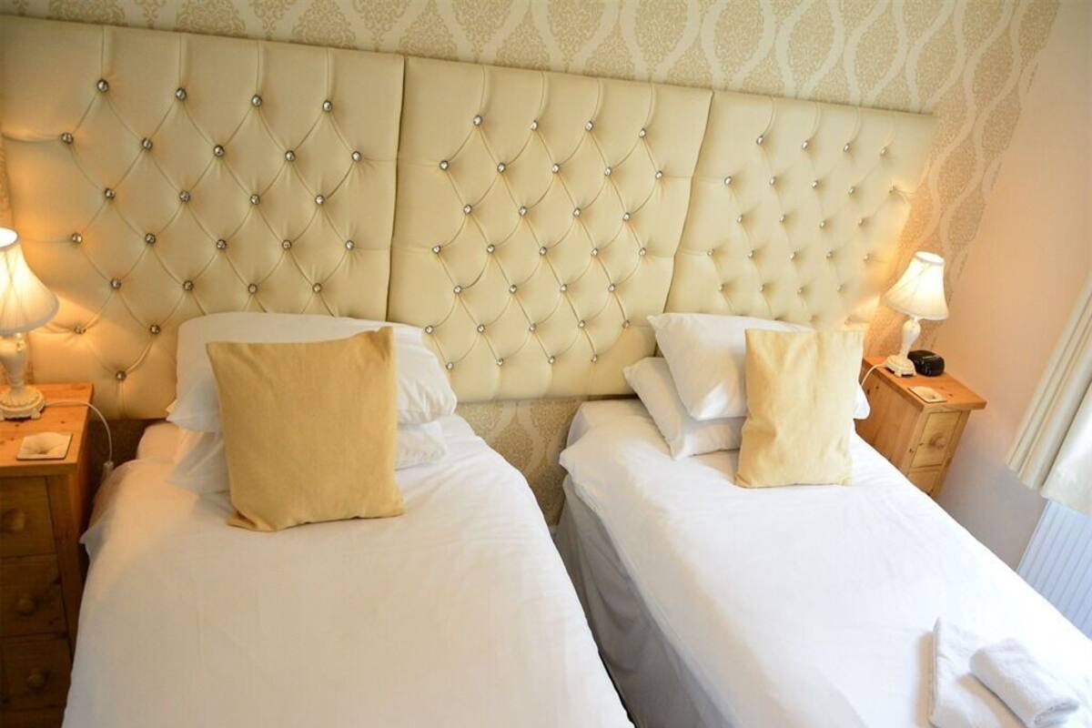 A hotel room with two beds, each with a tufted headboard and white linens. There are bedside tables with lamps and a small clock on one of them.