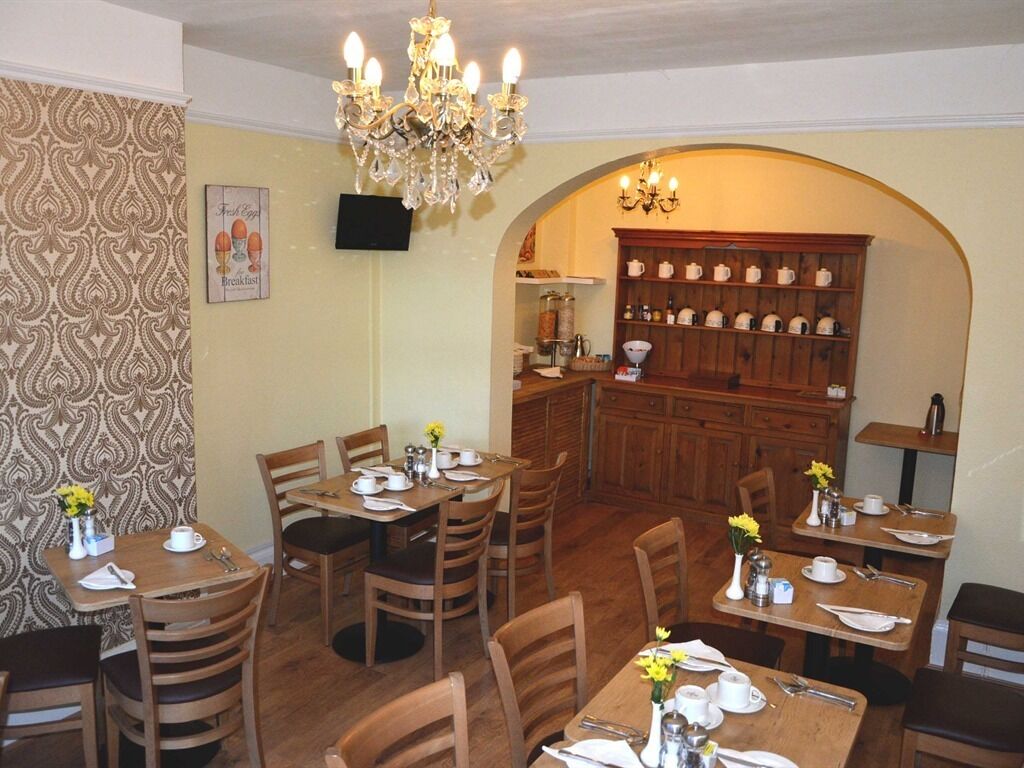 Una zona pranzo con tavoli e sedie in legno, un lampadario e una cucina visibile attraverso un arco.