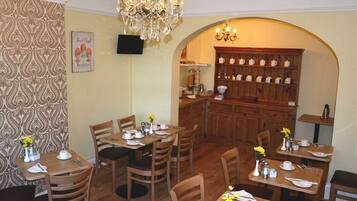 Una zona pranzo con tavoli e sedie in legno, un lampadario e una cucina visibile attraverso un arco.