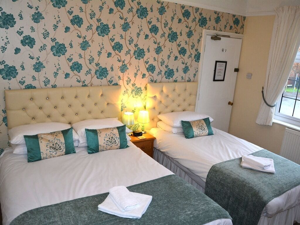 A hotel room with two beds, floral wallpaper, a door with a plaque, and a window with curtains.