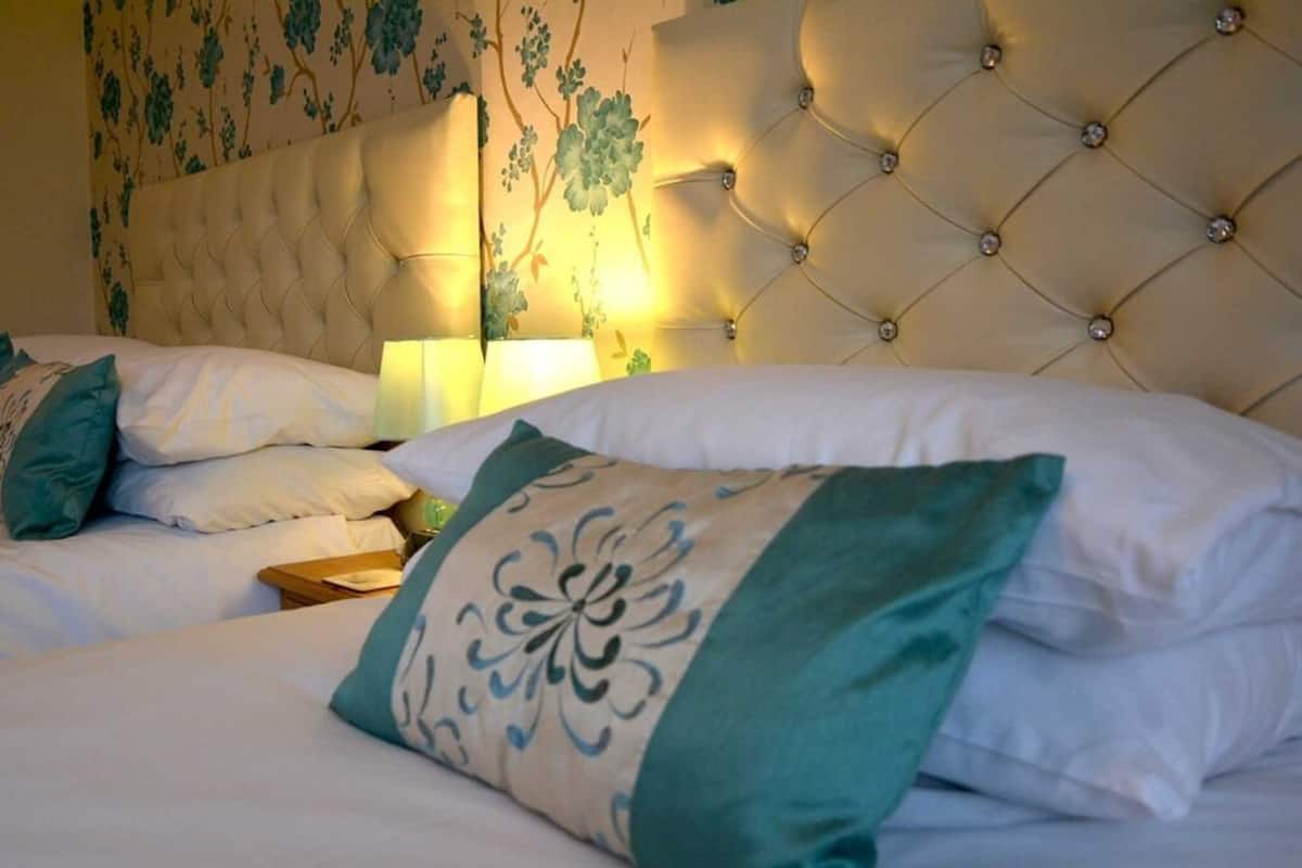 A hotel room with a tufted headboard, a bed with white and teal bedding, and a floral wallpaper design.