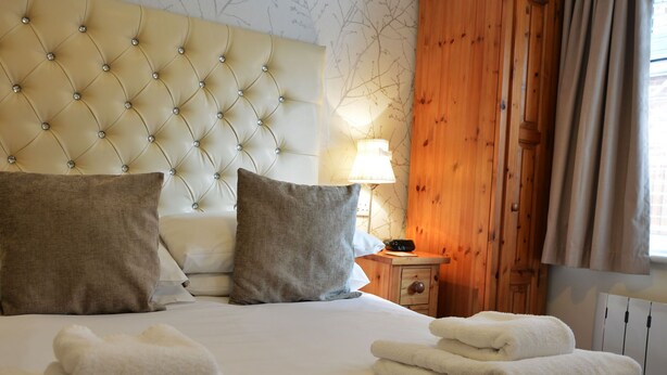 A neatly made bed with white linens and two pillows, a wooden nightstand with a lamp, and a wooden wardrobe. - Ashlea Guest House (Banbury)