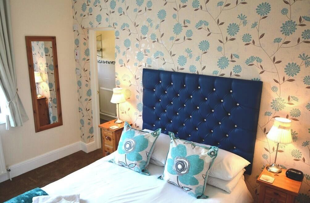Une chambre à coucher avec un grand lit, un papier peint à motifs floraux, une tête de lit capitonnée, deux tables de chevet avec des lampes et un miroir au mur.