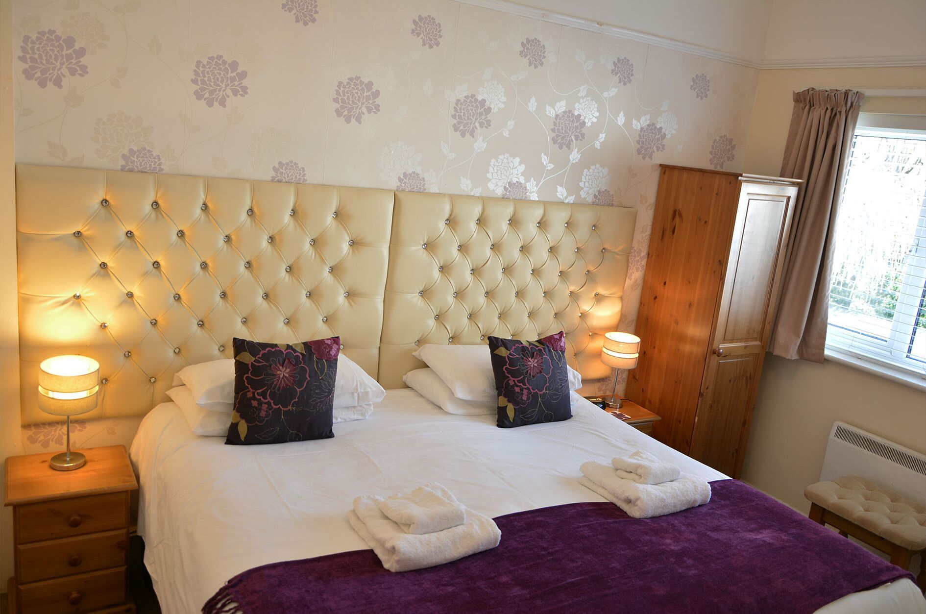 A hotel room with a tufted headboard, two bedside lamps, a bed with white linens, two decorative pillows, a purple throw blanket, and a window with curtains.