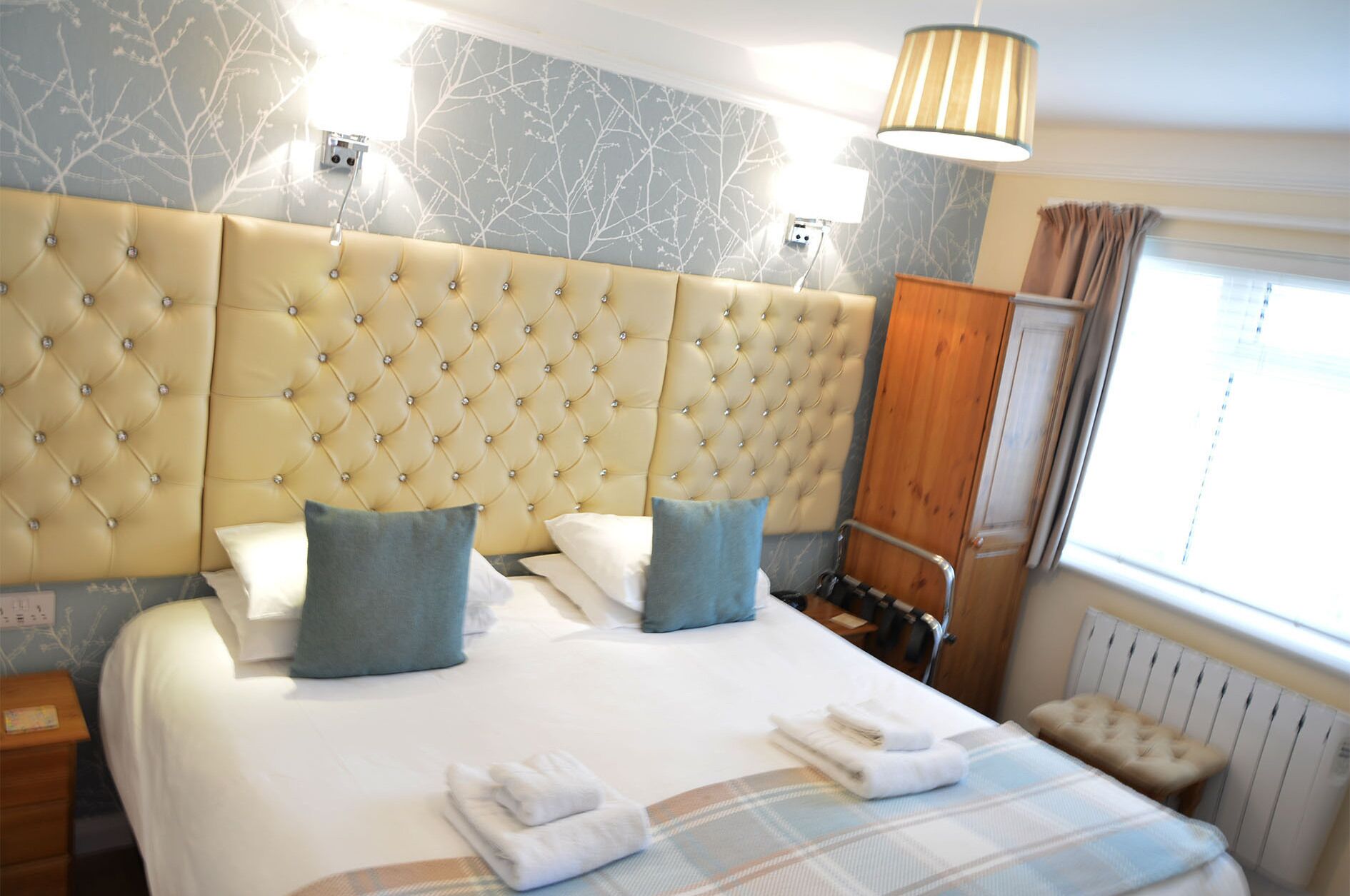 Habitación de hotel con una cama grande, dos almohadas, una mesita de noche, una lámpara y una ventana con cortinas.