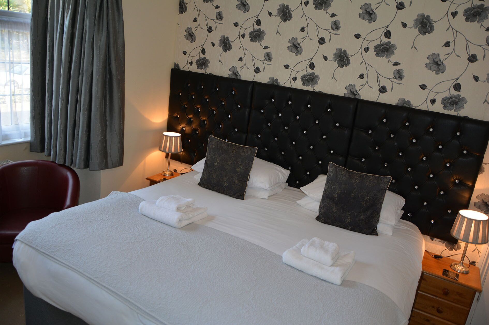 Een net opgemaakt bed met witte lakens en twee decoratieve kussens, een rode stoel en een nachtkastje met een lamp.