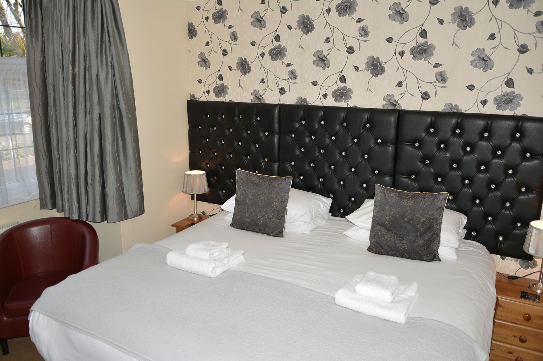 Een hotelkamer met een groot bed, een donker hoofdeinde, bloemenbehang, een rode stoel en een raam met gordijnen.