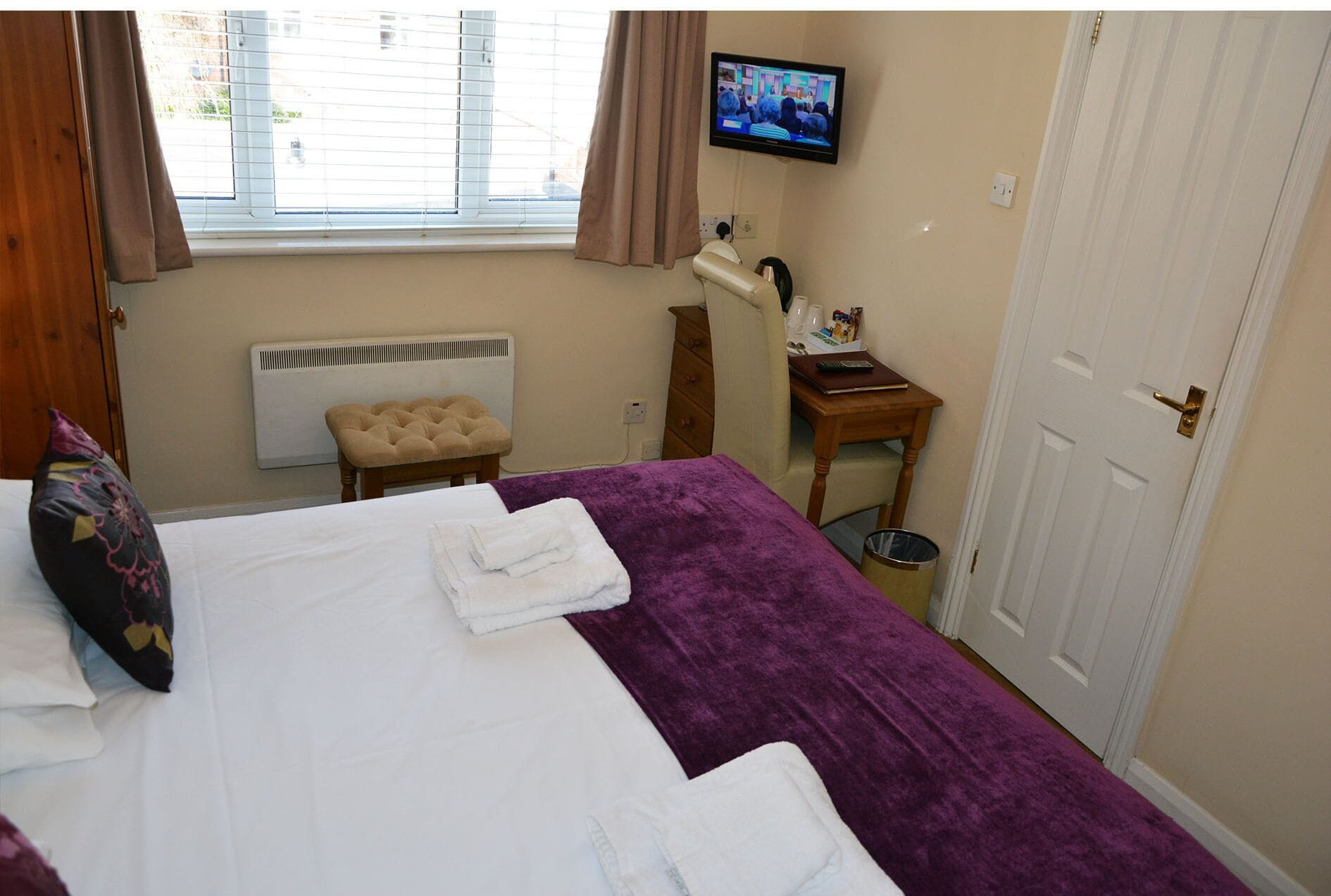 Habitación de hotel con cama, televisor, ventana con cortinas, radiador, una mesita con un mando a distancia y una puerta.