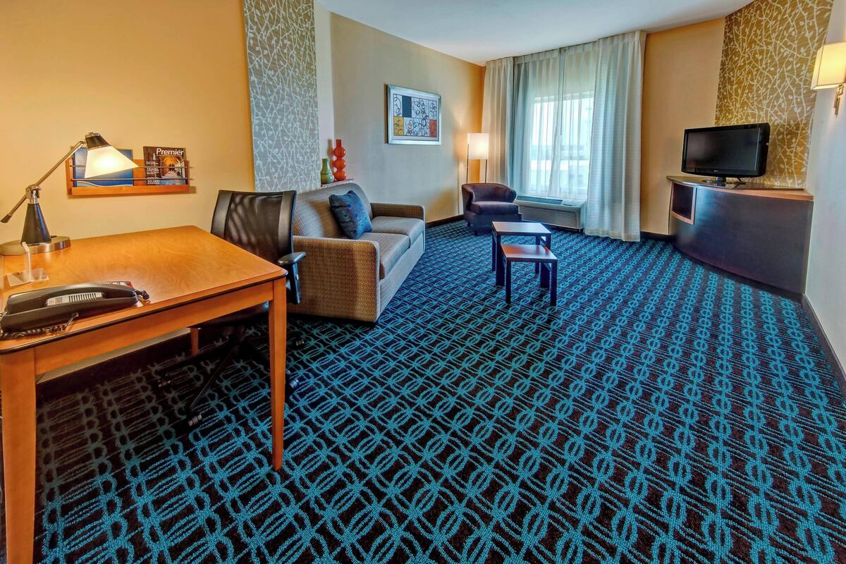 A hotel room with a patterned carpet, a sofa, a desk with a lamp, a television on a stand, and a window with curtains.