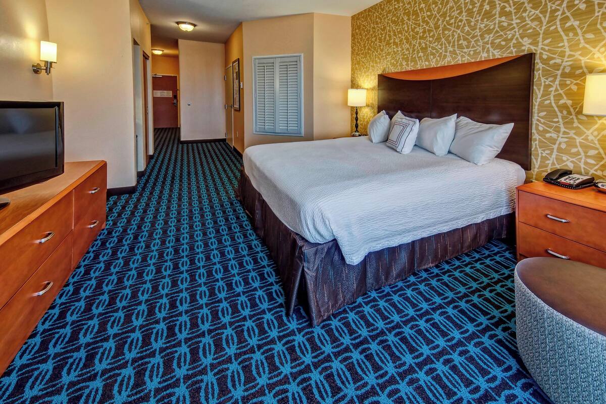 A hotel room with a large bed, a television, a desk with a lamp, and a patterned carpet.