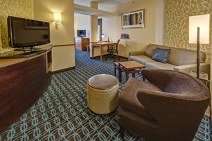 A hotel room with a flat-screen TV, a sofa, a chair, a small table, a lamp, and a patterned carpet.