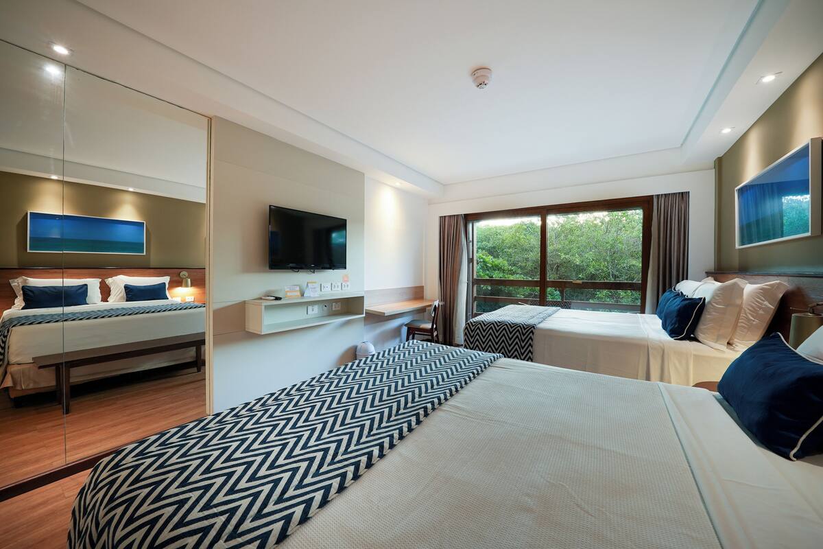 A modern hotel room with a large bed, a smaller bed, a TV, a window with curtains, and a mirror.