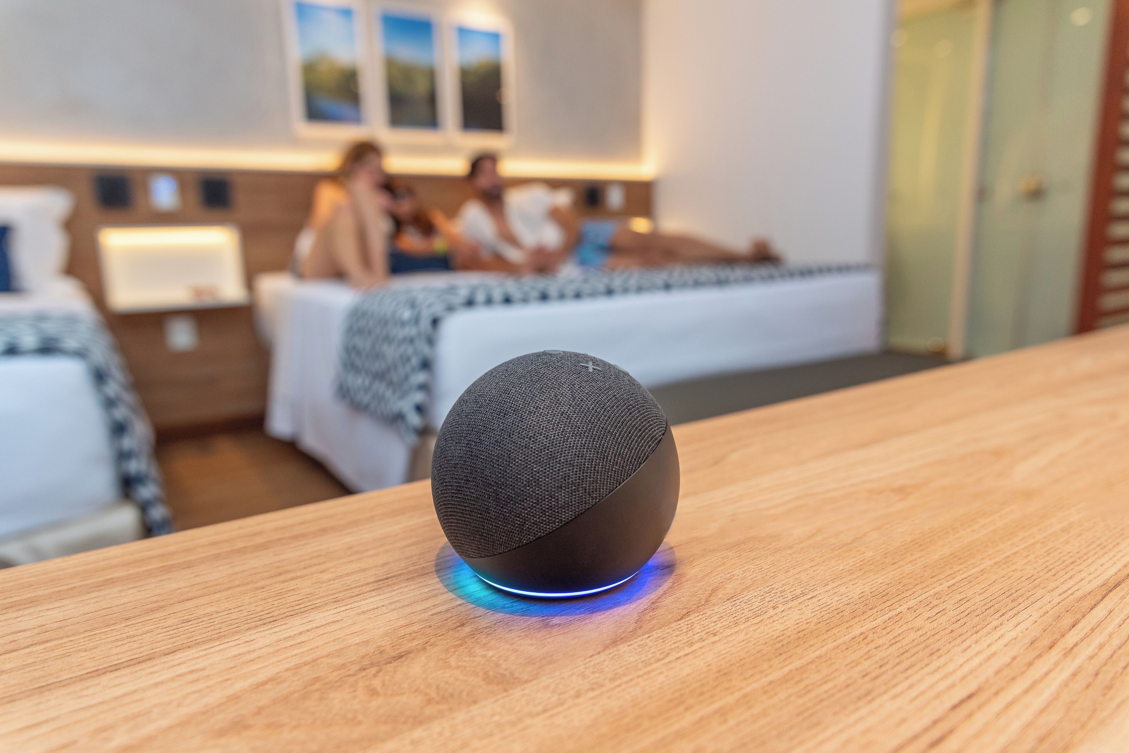 Una habitación de hotel moderna con un altavoz redondo cubierto de tela sobre una mesa de madera, una cama con una colcha estampada y una mesita de noche con una lámpara.