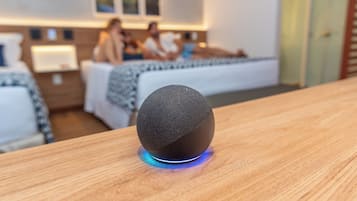 Una habitación de hotel moderna con un altavoz redondo cubierto de tela sobre una mesa de madera, una cama con una colcha estampada y una mesita de noche con una lámpara.