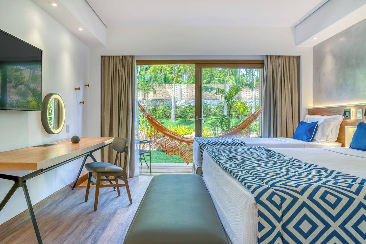 A hotel room with a bed, desk, chair, and a view of a garden through a glass door.