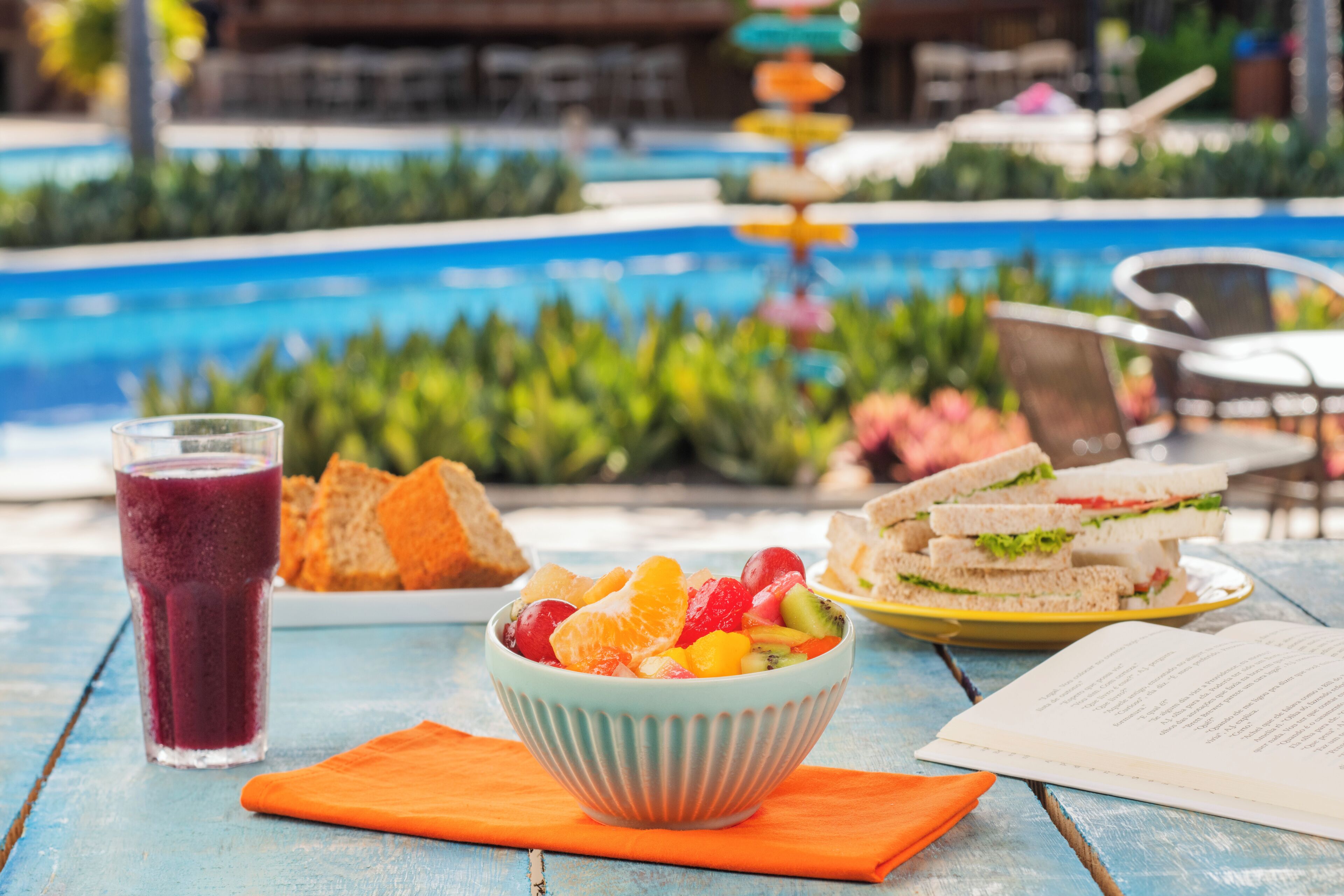 Una mesa con ensalada de frutas, sándwiches y un vaso de jugo, con una piscina al fondo.