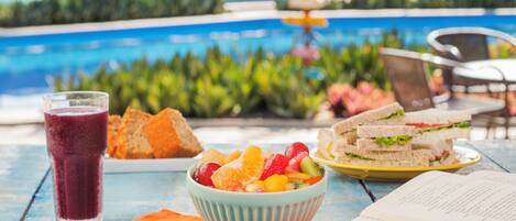Una mesa con ensalada de frutas, sándwiches y un vaso de jugo, con una piscina al fondo.
