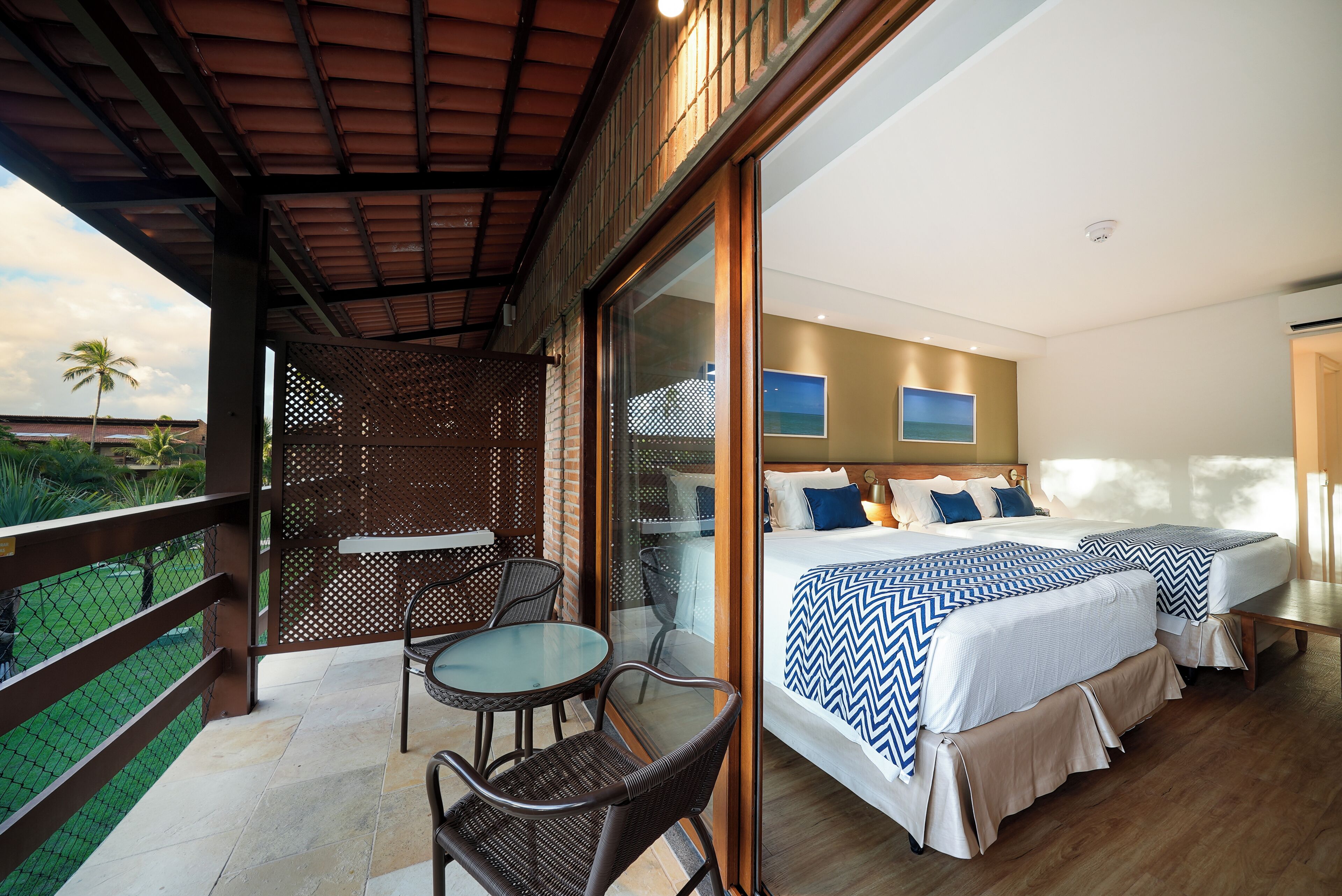 A balcony with a glass table and chairs, a bed with a blue and white chevron bedspread, and a view of palm trees.